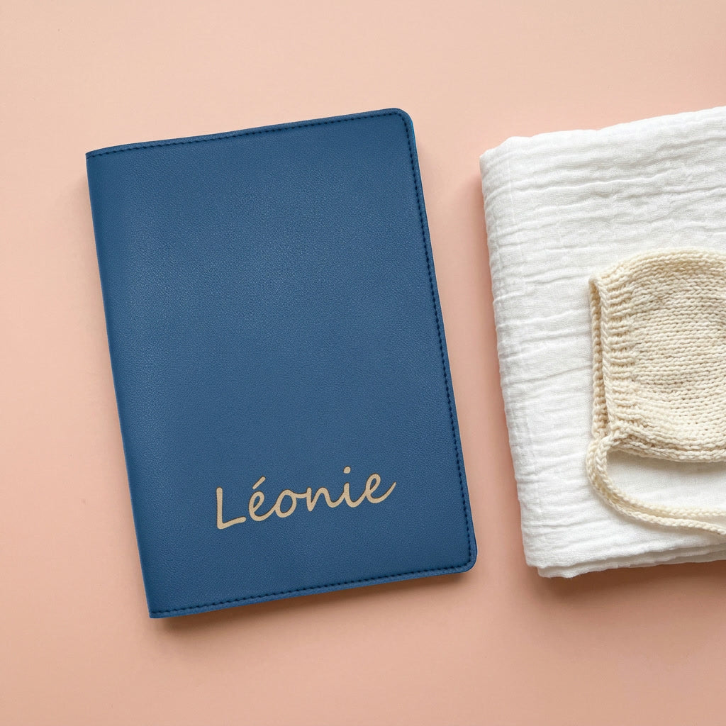 Mise en scène : protège carnet de santé bleu gravé « Léonie » près d'un bonnet de naissance