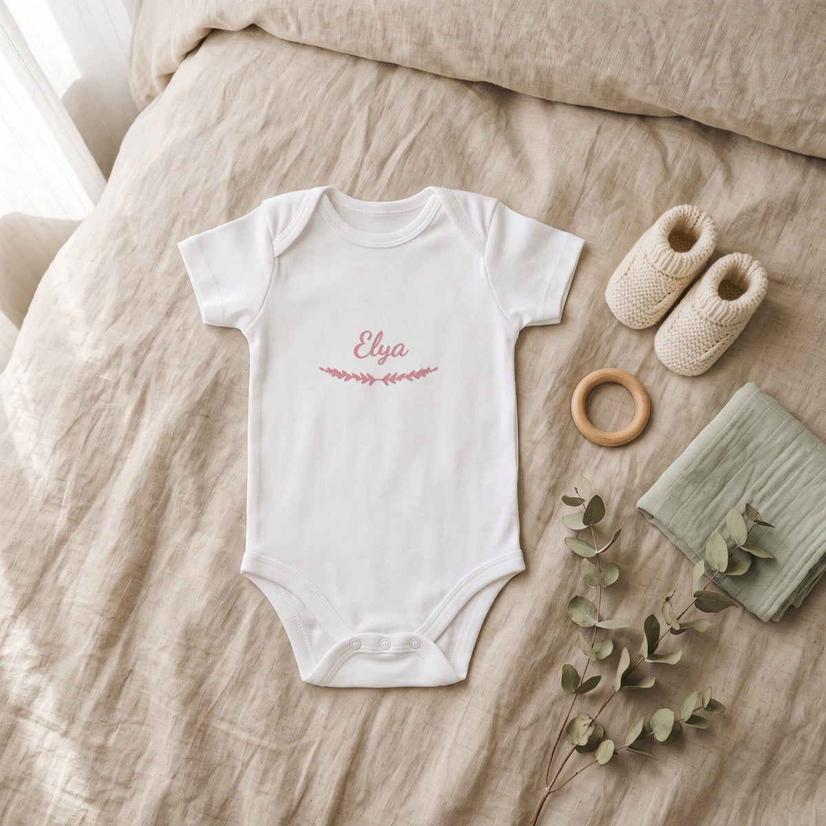 Body bébé blanc marqué « Elya » avec l'ornement Brindilles, posé à plat dans une chambre de bébé (visuel de mise en scène)