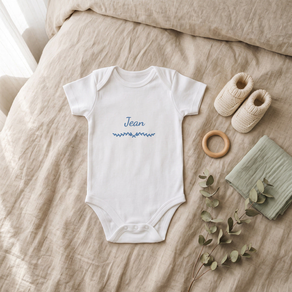 Body bébé blanc marqué « Jean » avec l'ornement Liane, posé à plat dans une chambre de bébé (visuel de mise en scène)