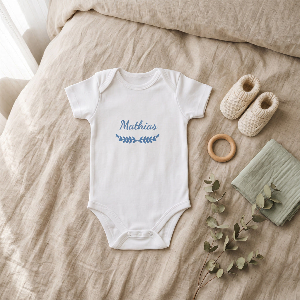 Body bébé blanc marqué « Mathias » avec l'ornement Feuillage, posé à plat dans une chambre de bébé (visuel de mise en scène)