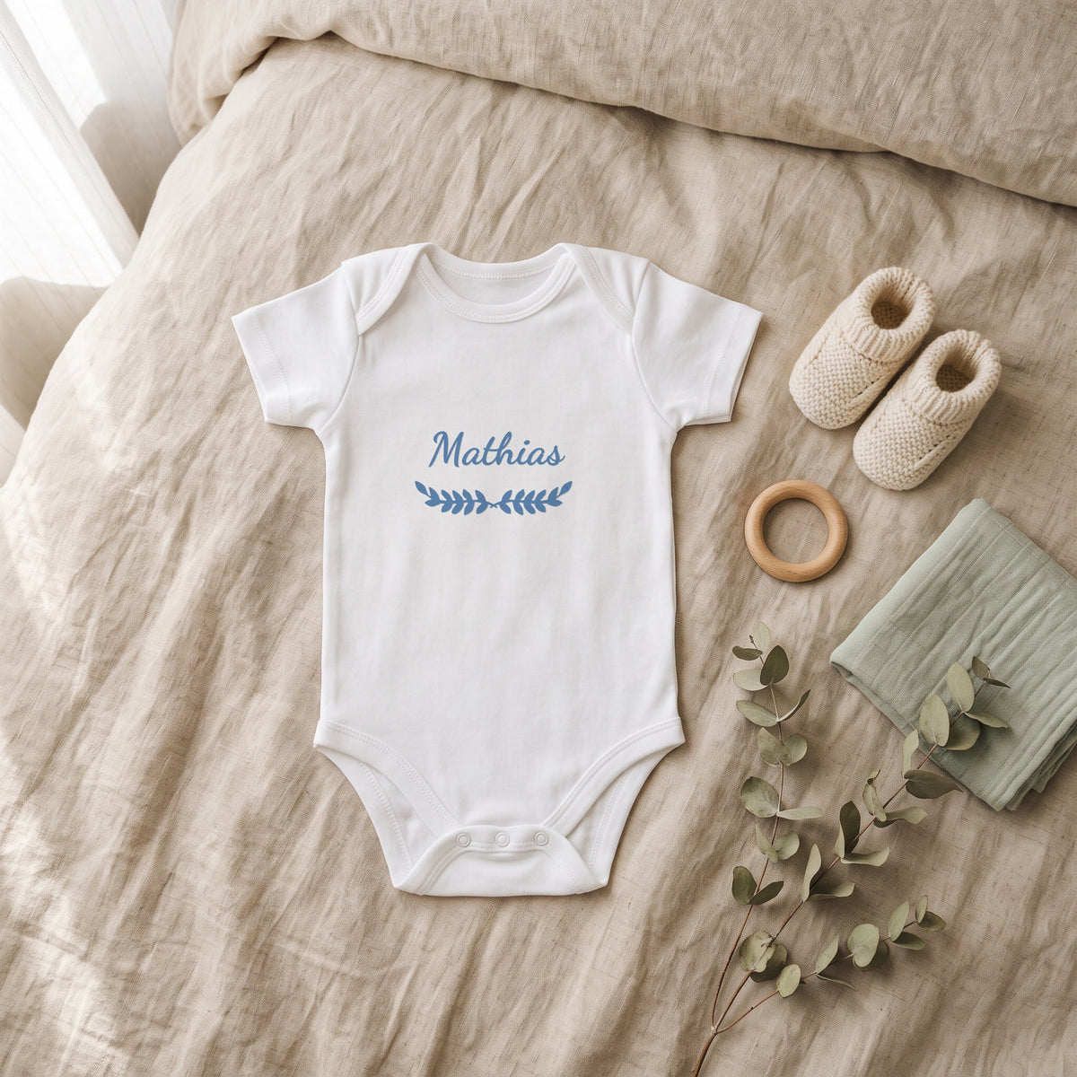 Body bébé blanc marqué « Mathias » avec l'ornement Feuillage, posé à plat dans une chambre de bébé (visuel de mise en scène)