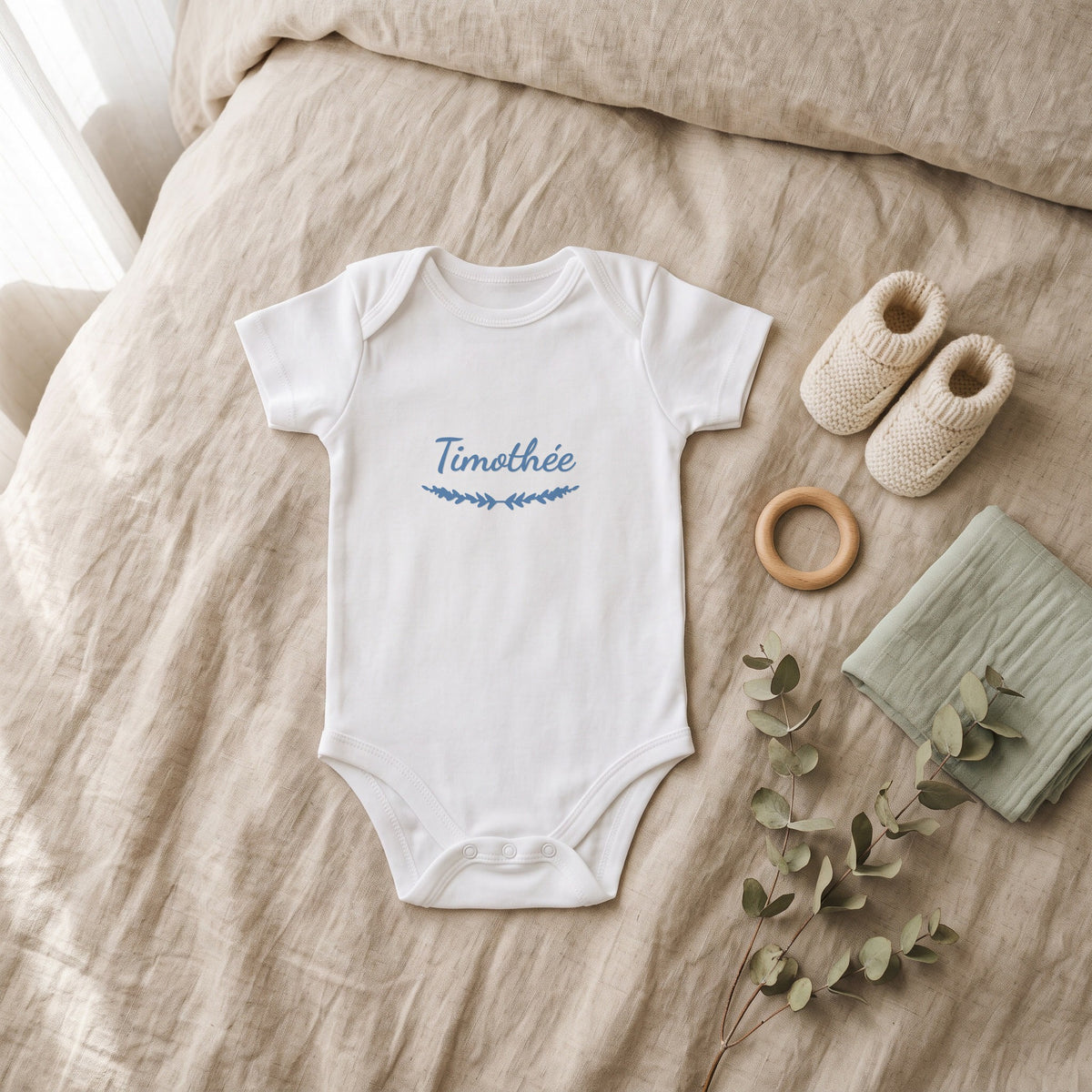 Body bébé blanc marqué « Timothée » avec l'ornement Brindilles, posé à plat dans une chambre de bébé (visuel de mise en scène)