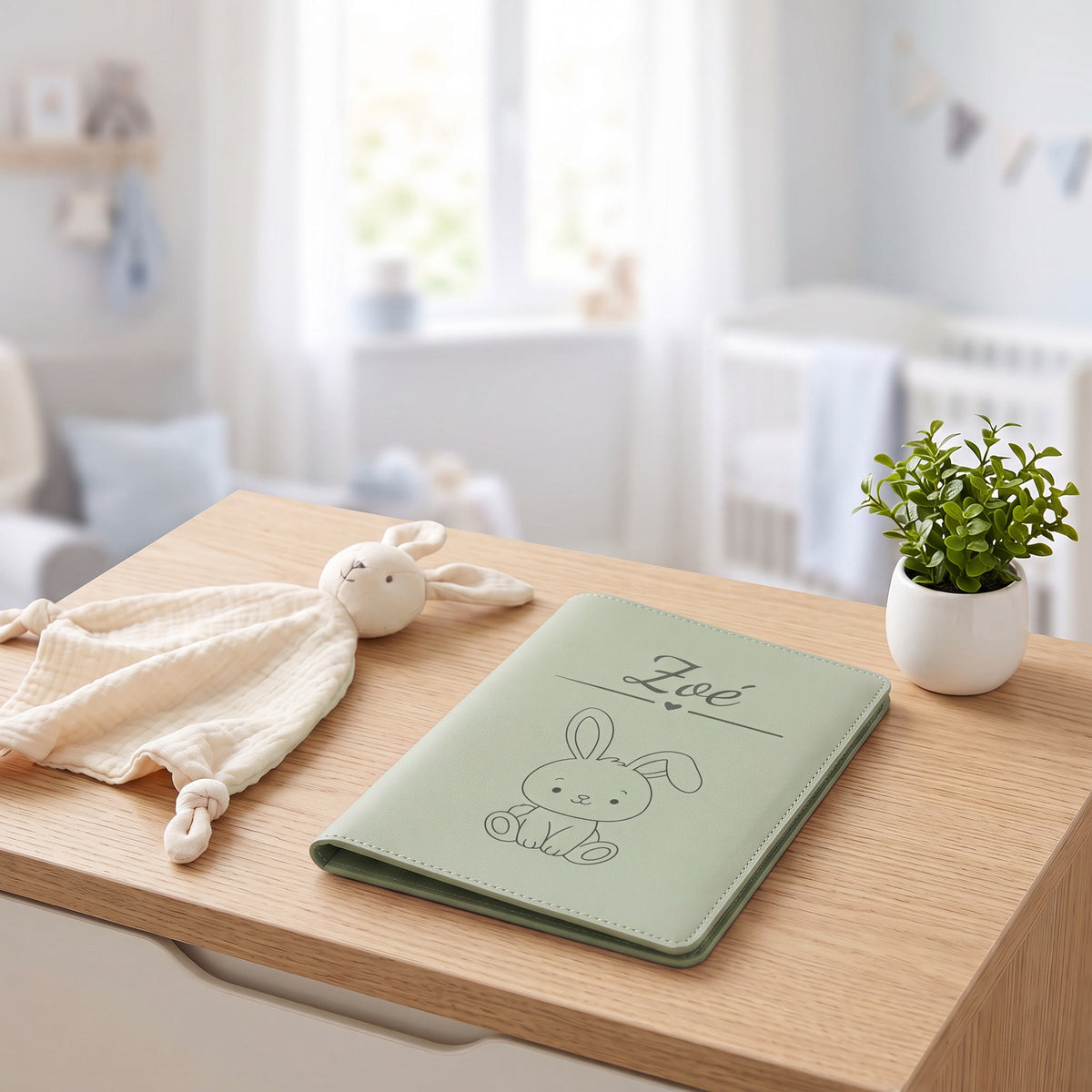 Protège carnet de santé bébé personnalisé – Poussin au foulard — fermé, exemple de gravure « Zoé » avec le motif lapin, sur une commode de chambre de bébé (visuel de mise en scène, coloris Vert sauge)
