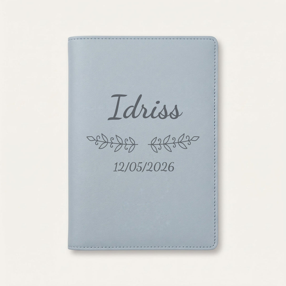 Protège carnet de santé personnalisé Idriss — Bleu nuage — prénom « Idriss », ornement Volutes et date de naissance « 12/05/2026 » gravés