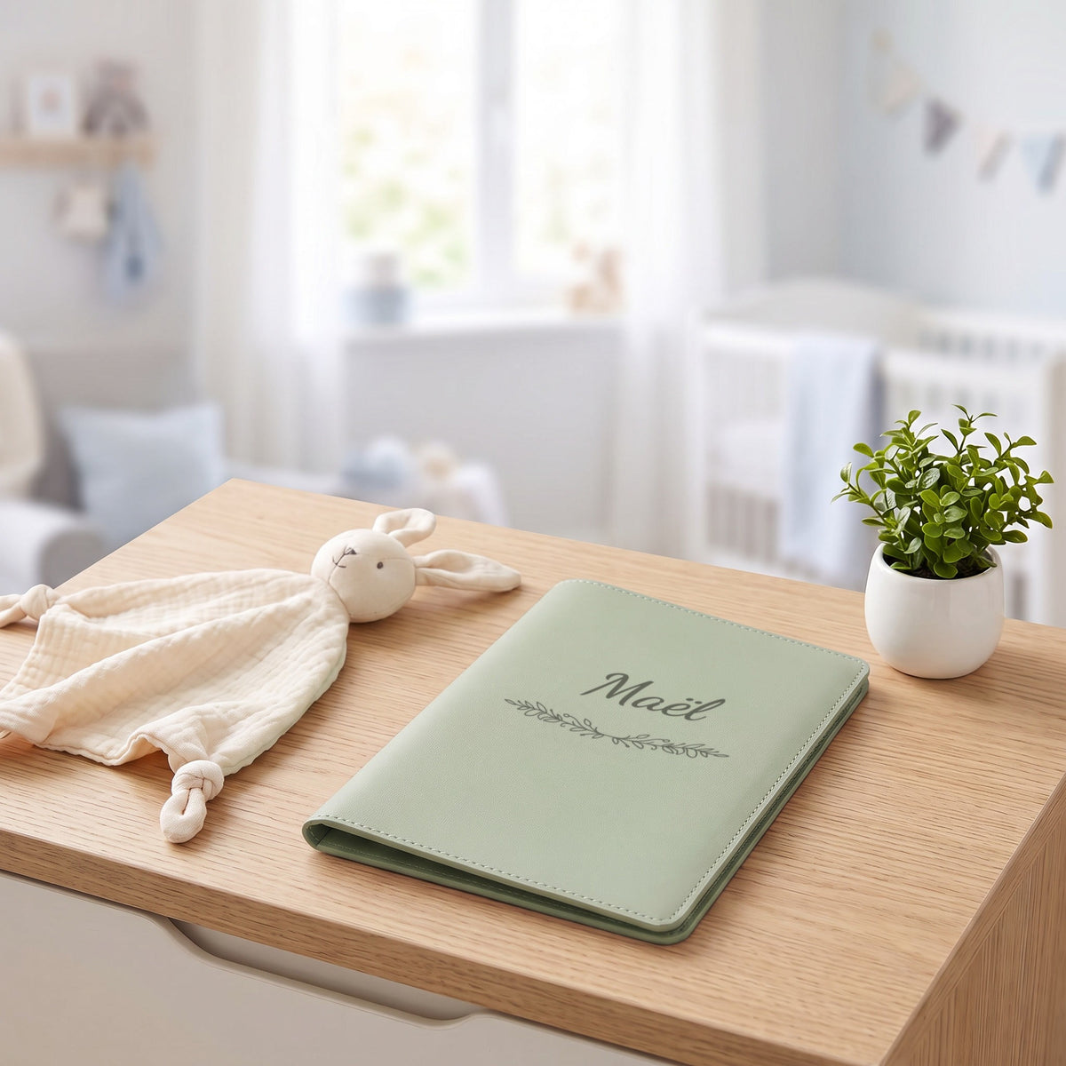 Protège carnet de santé personnalisé Maël — fermé, gravé « Maël » avec l'ornement Guirlande, sur une commode de chambre de bébé (visuel de mise en scène, coloris Vert sauge)